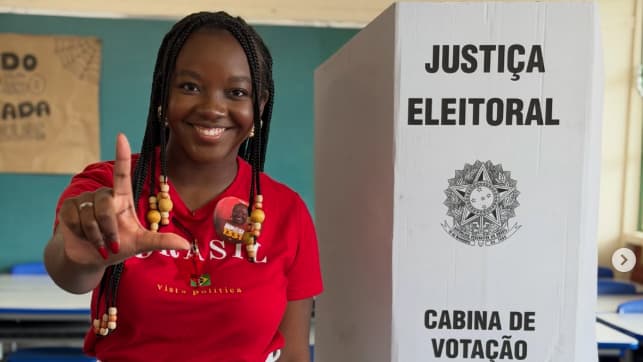 Ana Elisa posa junto a uma cabine de votação, com camiseta vermelha.