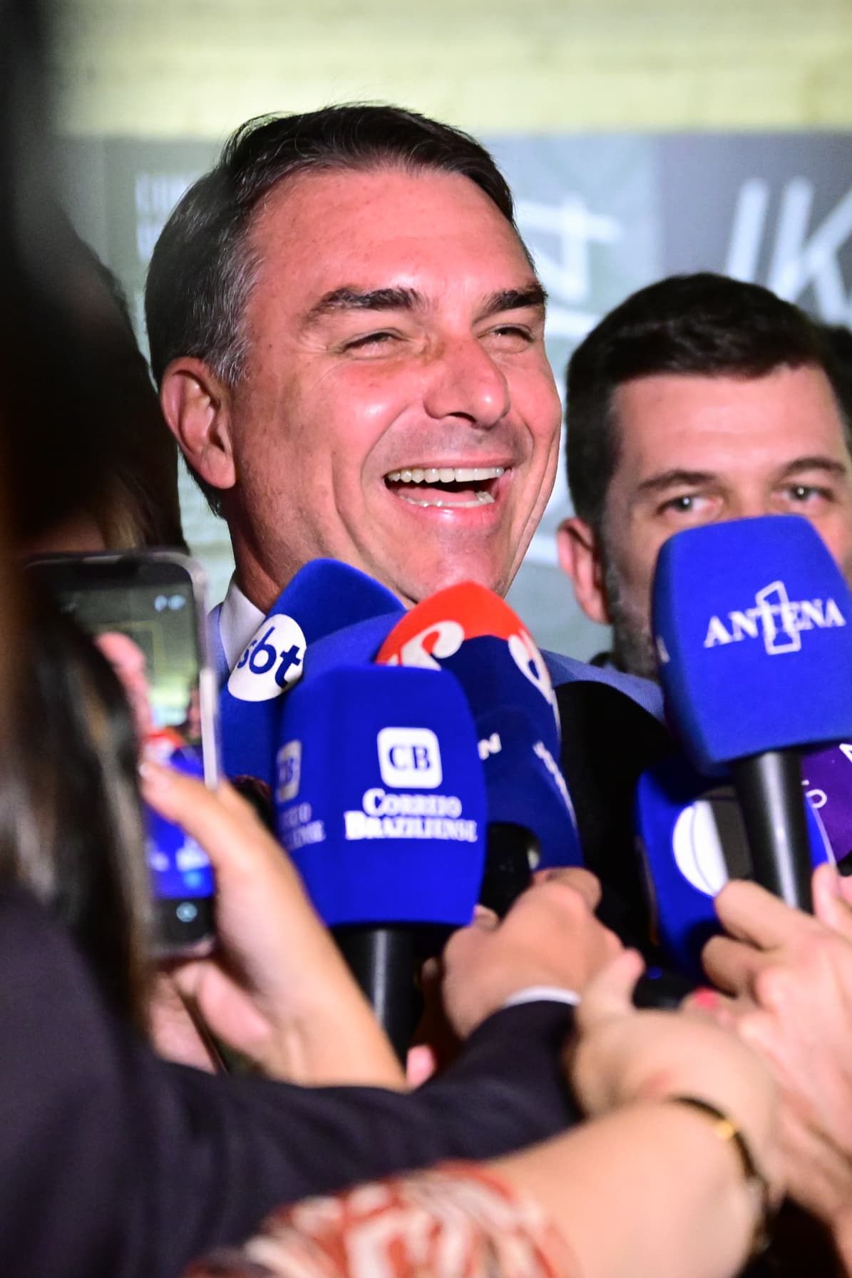Flávio Bolsonaro sorri durante entrevista cercado por microfones.