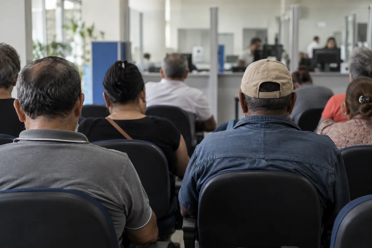 fila de pessoas de meia idade sentadas em cadeiras numa agência do INSS, fotografadas de costas.