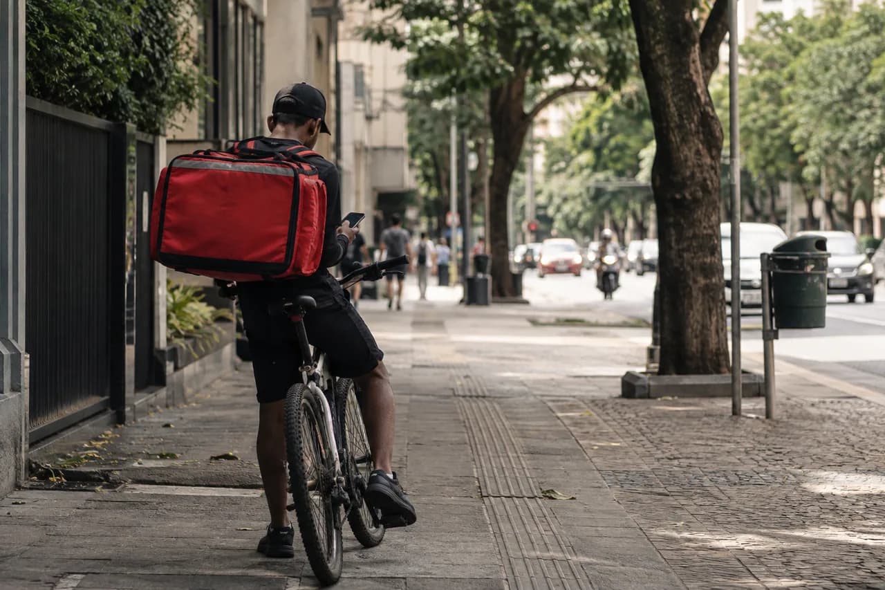 entregador de bicicleta esperando com o celular na mão em uma calçada, fotografado de costas.
