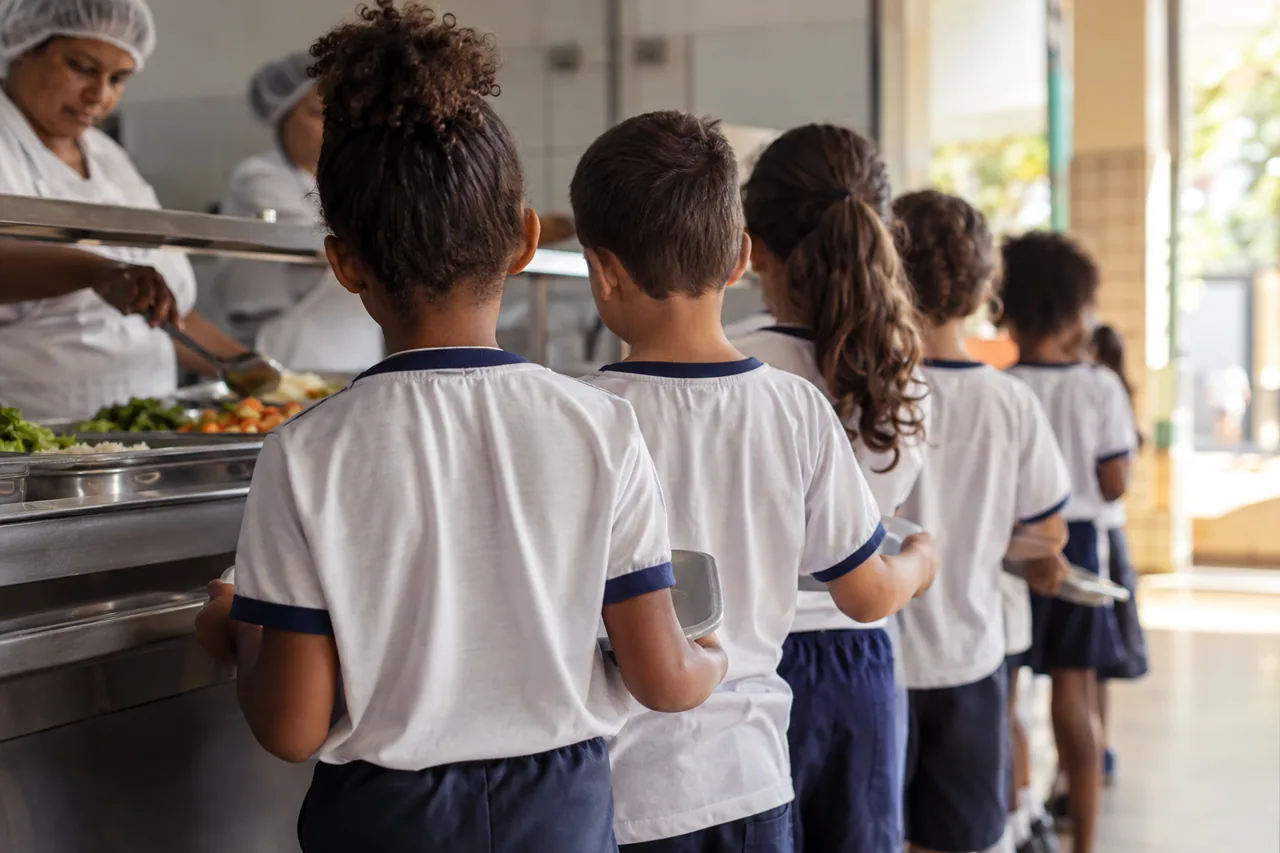 fila de crianças de uniforme escolar no refeitório, fotografadas de costas.