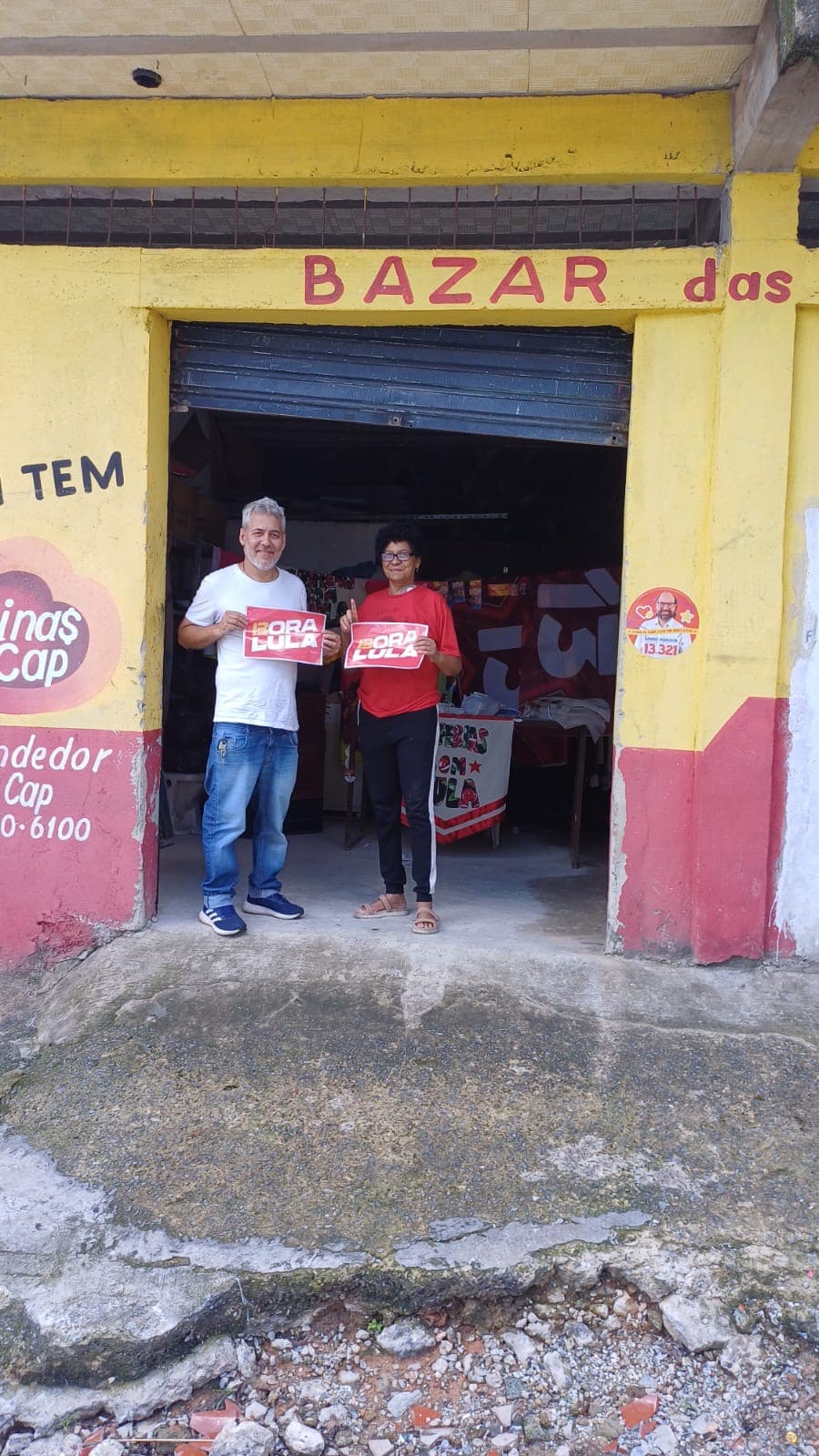 Duas pessoas exibem materiais Bora Lula em frente a estabelecimento