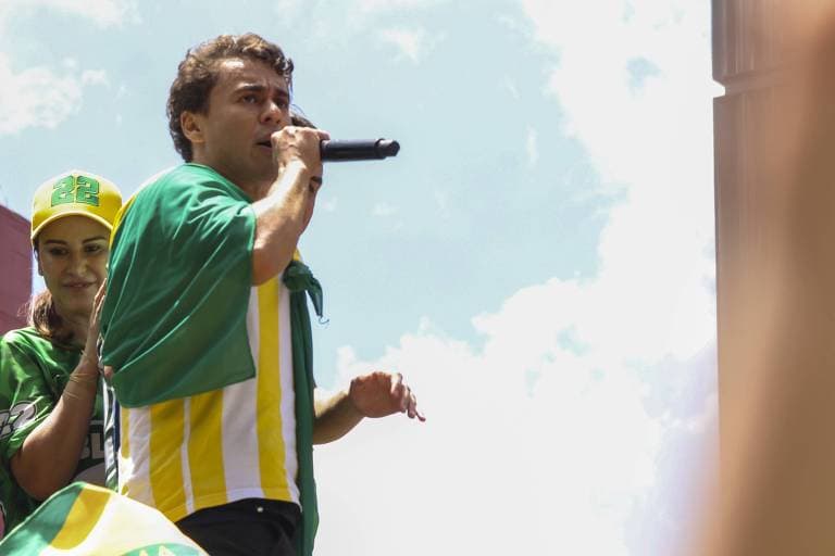 Nikolas Ferreira fala ao microfone em ato de apoio a Flávio Bolsonaro, em São Paulo, com camiseta nas cores do Brasil.
