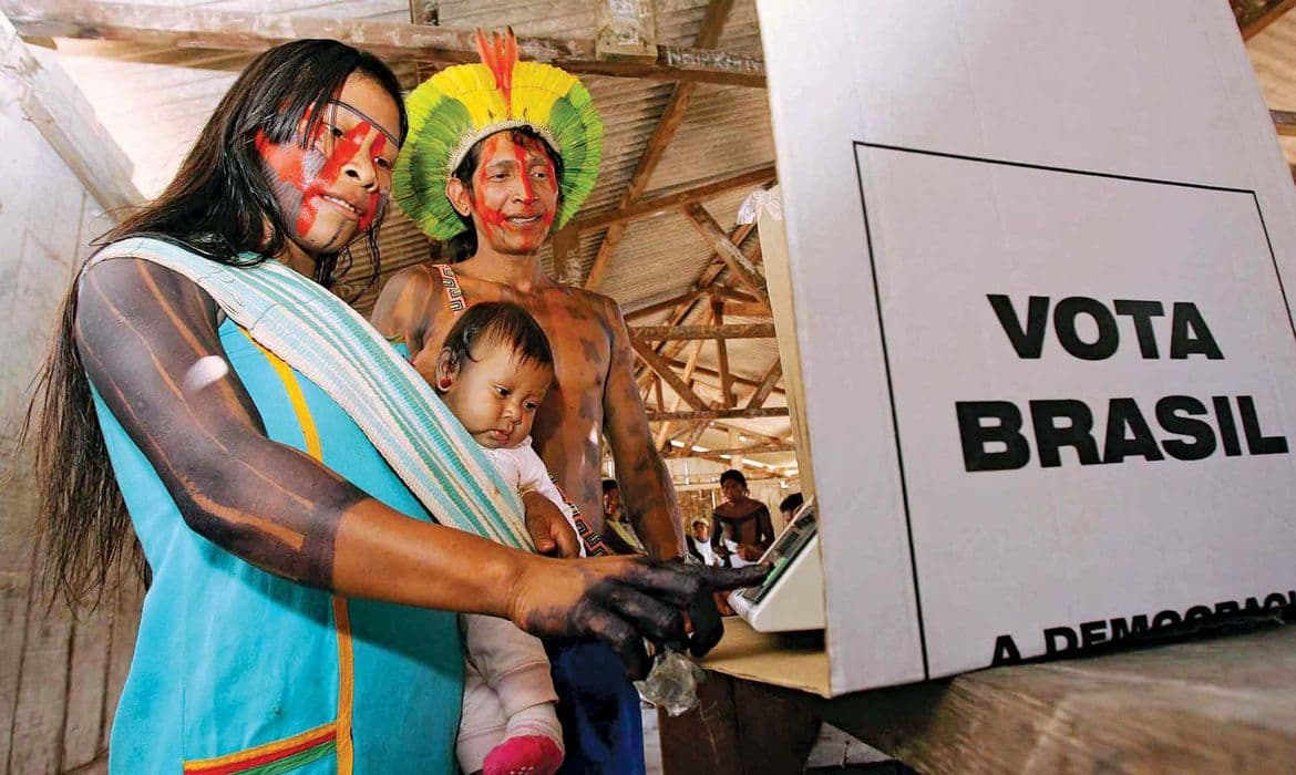 Eleitores indígenas junto a uma urna eletrônica, em fotografia de arquivo sobre participação eleitoral.
