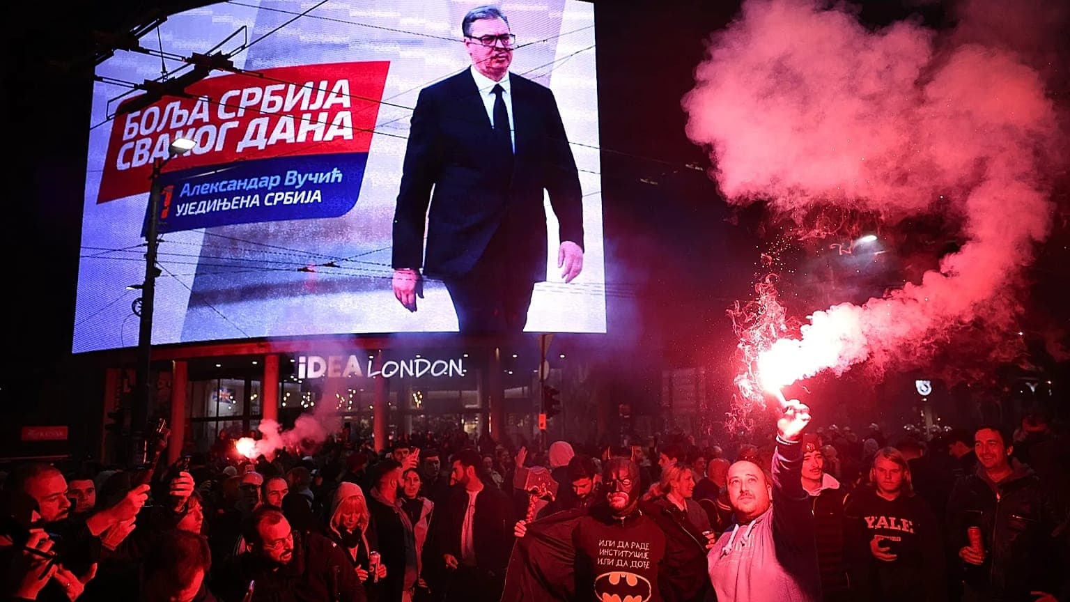 Pessoas celebram em Belgrado após Vučić reconhecer a derrota parlamentar, com a imagem do líder sérvio num painel urbano, em 25 de outubro de 2026.