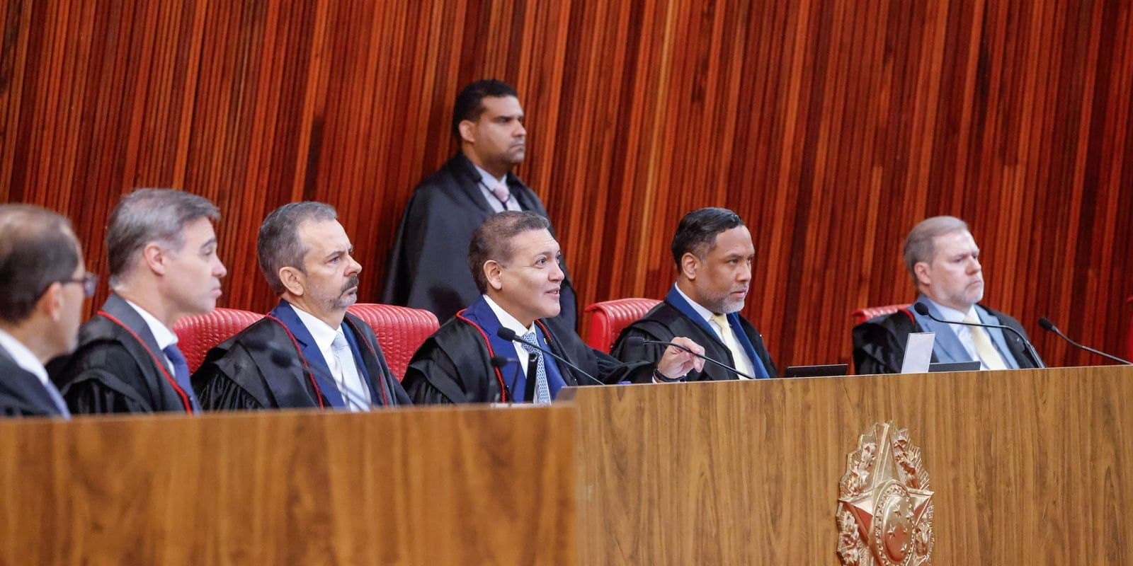 Integrantes de um tribunal, vestidos com togas, participam de sessão plenária em uma bancada com microfones.