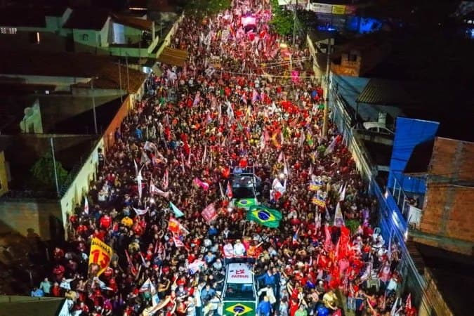 Vista aérea noturna de uma multidão em ato político, com bandeiras vermelhas e do Brasil, ocupando uma rua entre veículos e edificações.