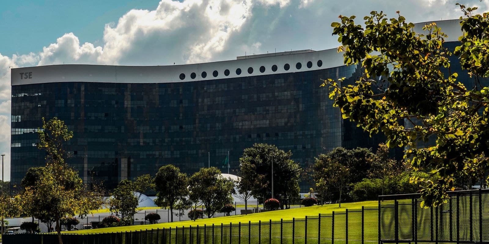 Vista externa do edifício-sede do Tribunal Superior Eleitoral, em Brasília, com árvores e área gramada em primeiro plano.