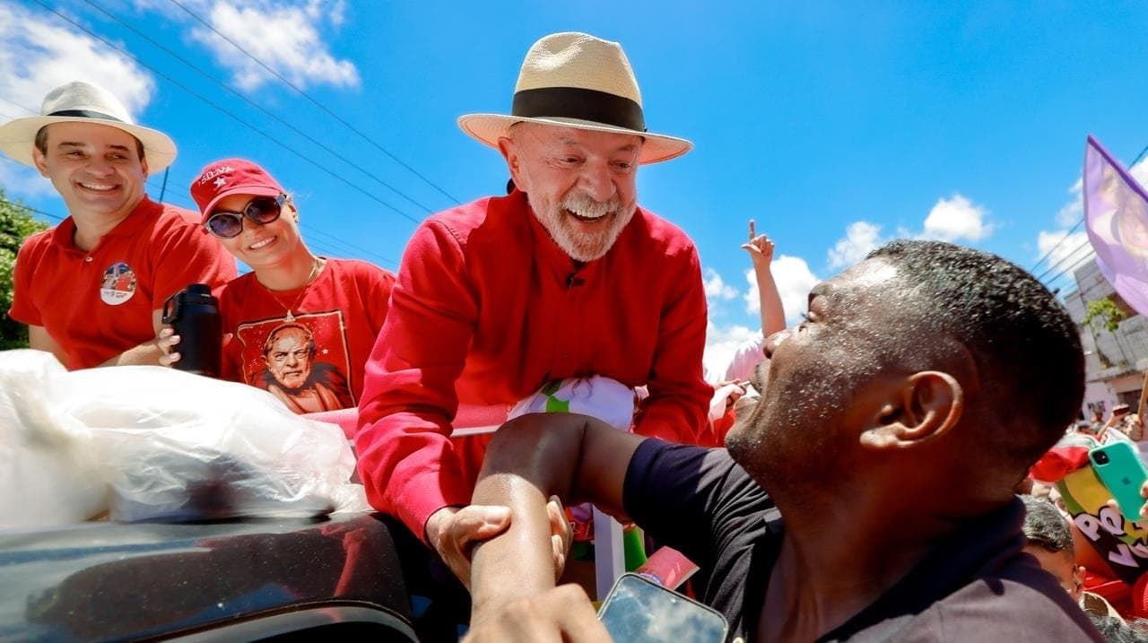 Homem de camisa vermelha e chapéu cumprimenta um apoiador em meio a um ato político ao ar livre.