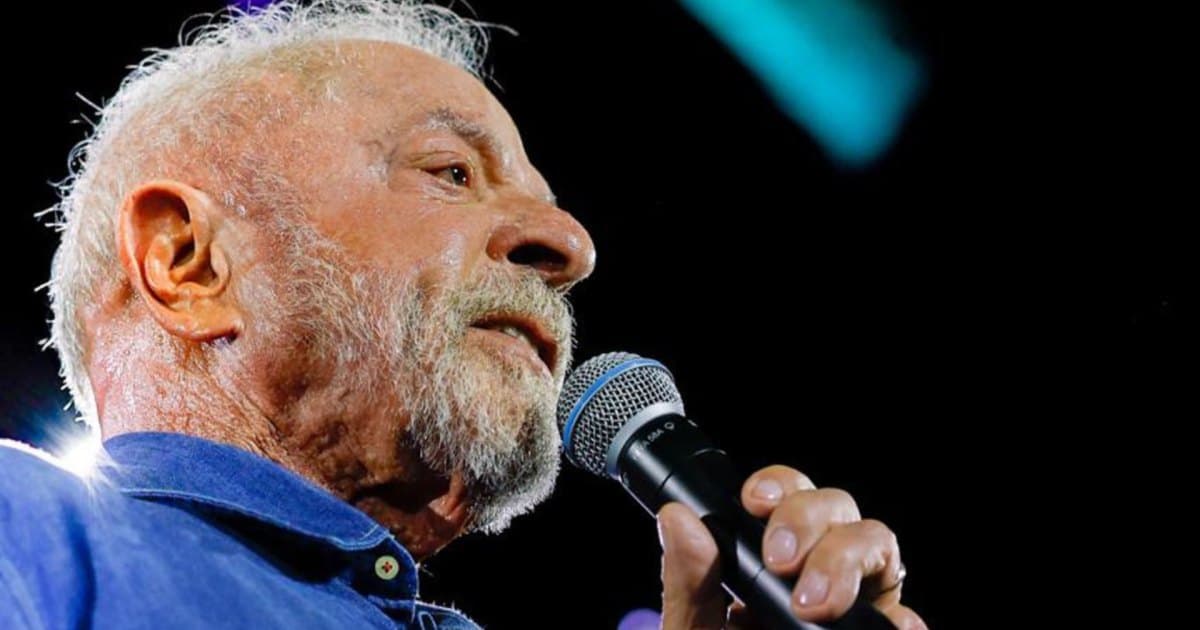 Homem idoso de barba branca segura um microfone durante evento público, em enquadramento fechado e visto de baixo.