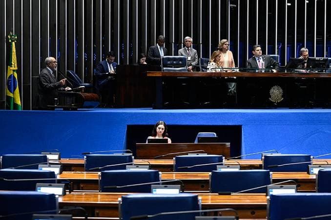 Vista do plenário do Senado, com parlamentares e servidores na mesa diretora e diversas cadeiras vazias.