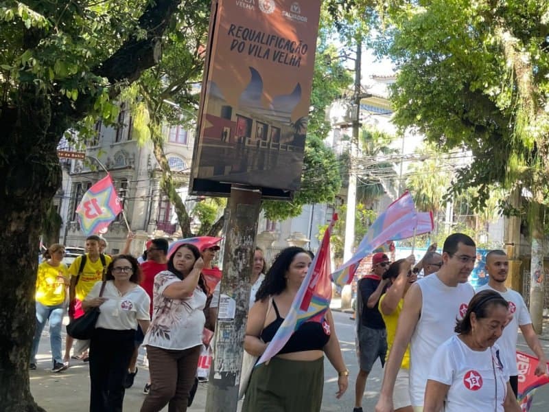 Grupo de manifestantes caminha por uma rua arborizada, carregando bandeiras coloridas e vestindo camisetas com o número 13.
