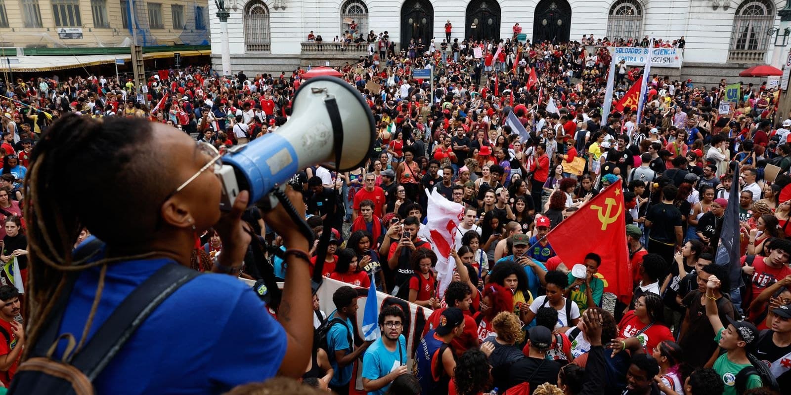 Jovem fala ao megafone em primeiro plano diante de uma grande multidão reunida em manifestação, com bandeiras e cartazes ao fundo.