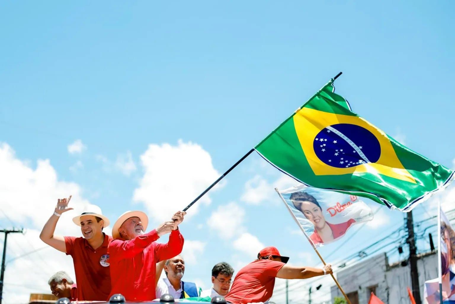 Homem de camisa vermelha agita uma bandeira do Brasil ao lado de apoiadores durante ato político ao ar livre.