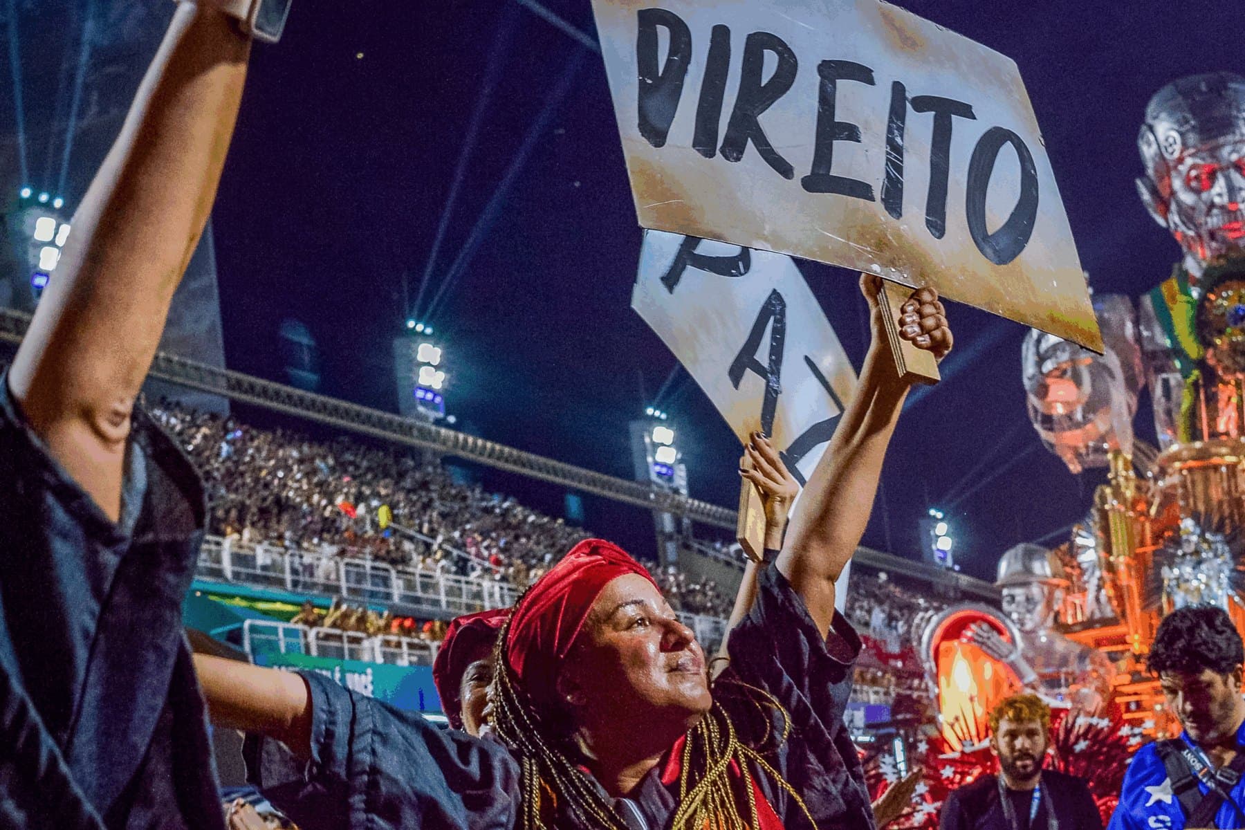 Integrantes de um desfile de carnaval erguem cartazes na Marquês de Sapucaí, diante das arquibancadas lotadas.