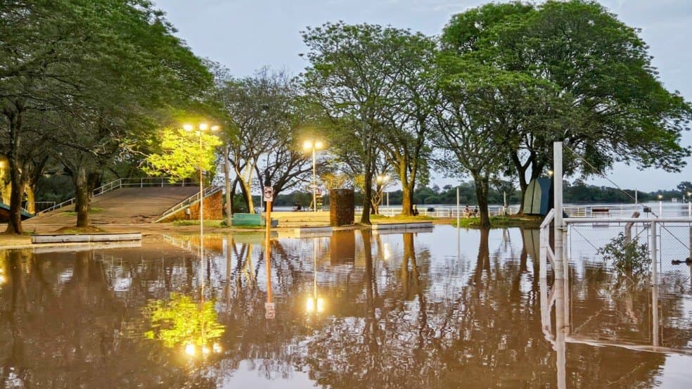 Área pública arborizada alagada, com água cobrindo caminhos e refletindo árvores, postes acesos, cercas e estruturas do local.